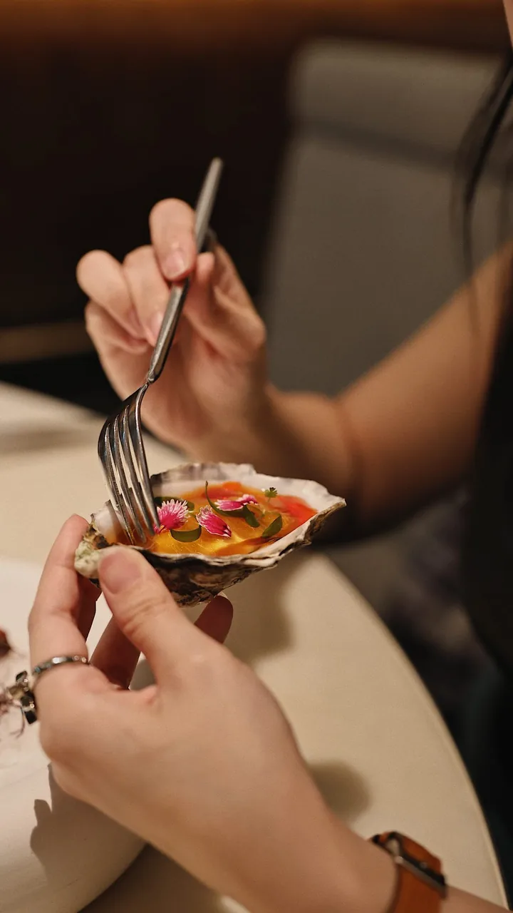 雙手托著盛裝料理的貝殼，一把叉子靠近帶有小花與葉片的食物。