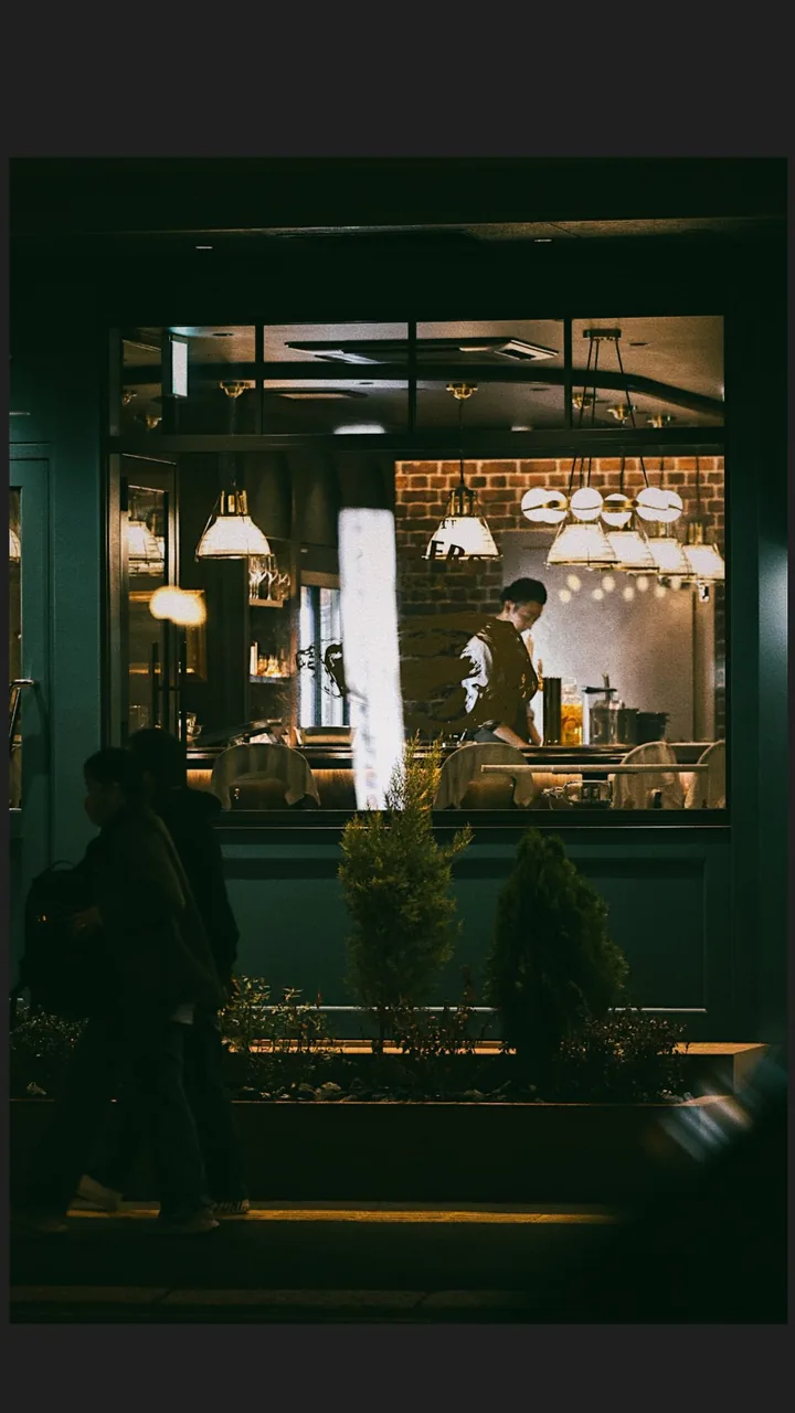 從街道望向深綠色店面窗內，暖色吊燈照著桌椅與一名成人。